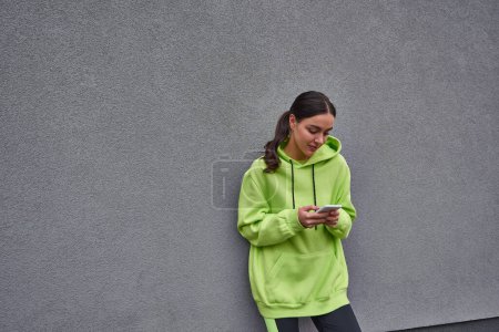 junge Frau in lindfarbenem Kapuzenpulli und Leggings mit Smartphone in der Nähe einer grauen Betonwand