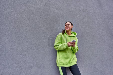 mujer feliz en color lima sudadera con capucha y leggings mensajería en el teléfono inteligente cerca de pared de hormigón gris