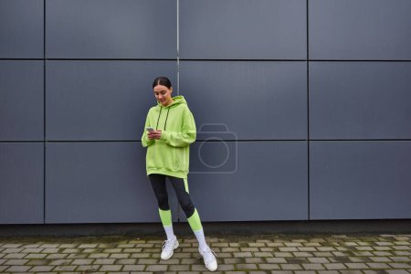 mujer feliz en color lima con capucha y leggings mensajería en el teléfono inteligente cerca de la pared gris al aire libre