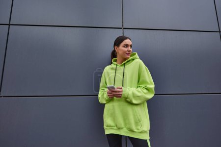 mujer alegre en color lima sudadera con capucha que sostiene el teléfono inteligente y mirando hacia otro lado mientras está de pie cerca de la pared gris