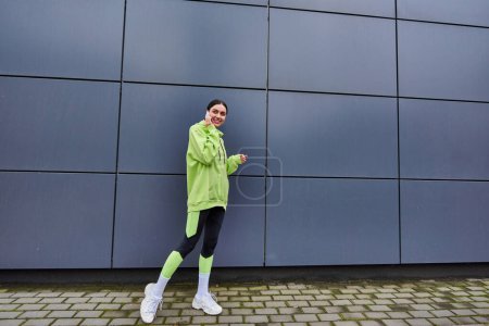 happy woman in lime color hoodie and leggings talking on smartphone near grey wall outdoors
