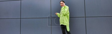 mujer feliz en color lima sudadera con capucha y leggings hablando en el teléfono inteligente cerca de la pared gris al aire libre, pancarta