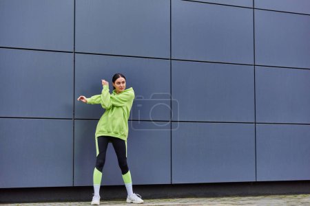 sportswoman in hoodie and leggings working out near grey wall outdoors, fresh air and sport