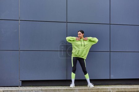 joven deportista con capucha y polainas calentándose cerca de la pared gris al aire libre, aire fresco y deporte