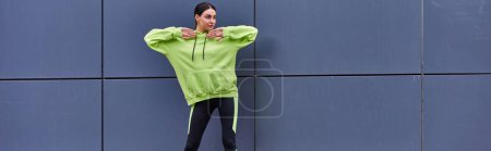 Sportlerin in Kapuzenpulli und Leggings wärmt sich an grauer Wand im Freien auf, frische Luft und Sportbanner