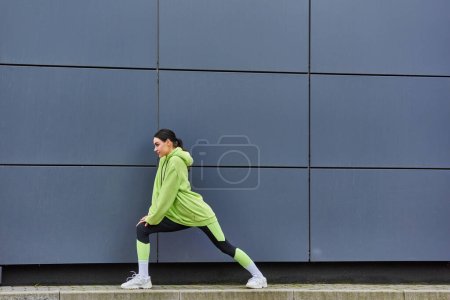 joven deportista con capucha y polainas que estira las piernas cerca de la pared gris al aire libre, la energía y el deporte