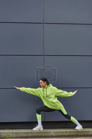 joven deportista en ropa deportiva con las manos extendidas estirando las piernas cerca de la pared gris al aire libre