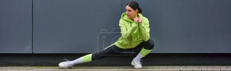 young sportswoman in hoodie and leggings doing lunges near grey wall outdoors, sport banner