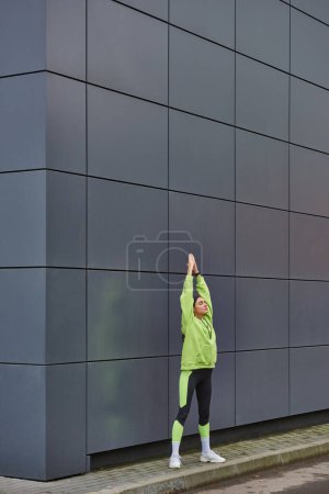 joven atlética en sudadera con capucha y polainas calentándose cerca de la pared gris al aire libre, motivación y deporte
