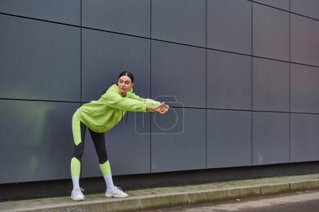 mujer atlética con capucha y polainas calentándose cerca de la pared gris al aire libre, motivación y deporte
