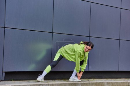 mujer enérgica con capucha y polainas calentándose cerca de la pared gris al aire libre, motivación y deporte