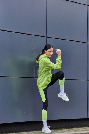 athletische Läuferin in Kapuzenpulli und Leggings beim Aufwärmen an der grauen Wand im Freien, Motivation