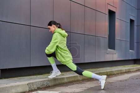athletische Läuferin in Kapuzenpulli und Leggings beim Ausfallschritt nahe grauer Wand im Freien, Motivation