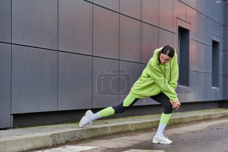 mujer atlética en sudadera con capucha color lima y polainas haciendo embestidas cerca de la pared gris al aire libre, motivación