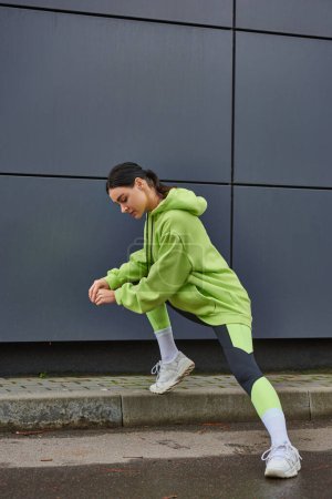athletische Frau in lindfarbenem Kapuzenpulli und Leggings, die sich in der Nähe einer grauen Wand im Freien ausstrecken, Motivation