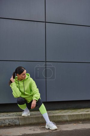 sportswoman in lime color hoodie and leggings stretching and sitting near grey wall outdoors, sport