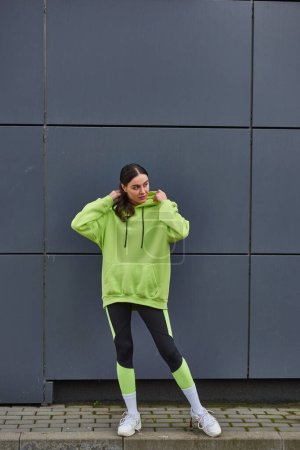 attraktive Sportlerin in kalkfarbenem Kapuzenpulli und Leggings, die neben grauer Wand im Freien steht, Sport