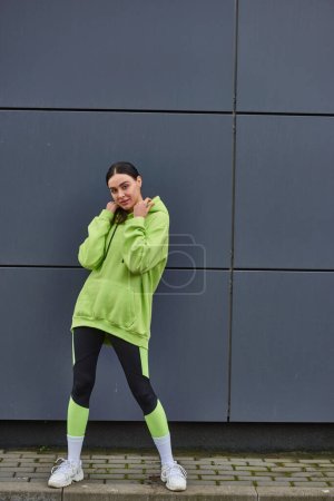 beautiful sportswoman in lime color hoodie and leggings standing near grey wall outdoors, sport