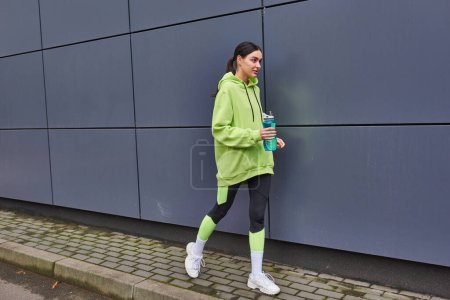 attraktive Sportlerin in Kapuzenpulli und Leggings, die mit Wasserflasche in der Nähe der grauen Wand im Freien spaziert