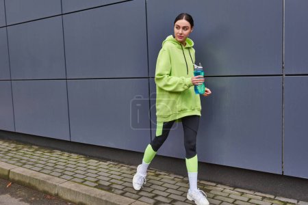 schöne Sportlerin in Kapuzenpulli und Leggings, die mit einer Flasche Wasser in der Nähe der grauen Wand im Freien spazieren geht