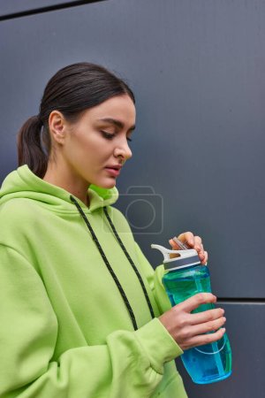 junge brünette Sportlerin in lindfarbenem Kapuzenpullover mit Wasserflasche nahe grauer Wand im Freien