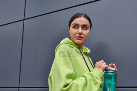junge attraktive Sportlerin in kalkfarbenem Kapuzenpulli mit Wasserflasche an der grauen Wand im Freien