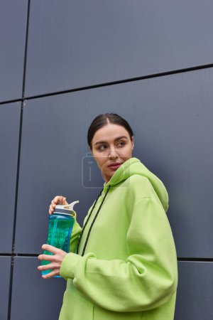 junge Sportlerin in lindfarbenem Kapuzenpulli mit Wasserflasche in der Nähe der grauen Wand im Freien, schaut weg