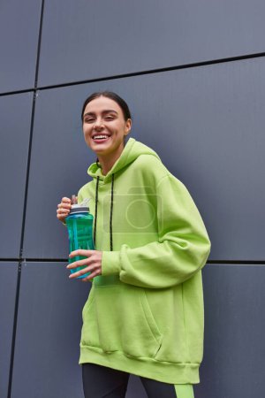 fröhliche und junge Sportlerin in kalkfarbenem Kapuzenpulli mit Wasserflasche an der grauen Wand im Freien