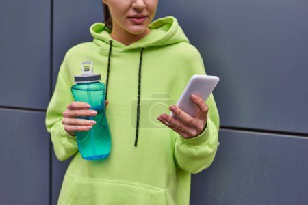 cropped sportswoman in hoodie holding bottle of water and using smartphone near grey wall outdoors