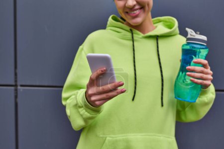 femme sportive cultivée et heureuse à capuche tenant une bouteille d'eau et utilisant un smartphone à l'extérieur