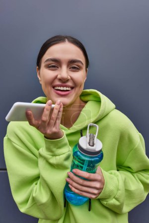 fröhliche Sportlerin im Kapuzenpullover mit Wasserflasche und Audiobotschaft auf dem Smartphone