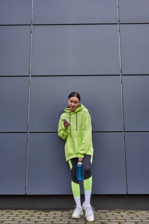 jolie sportive en sweat à capuche tenant bouteille d'eau et utilisant smartphone près du mur gris à l'extérieur