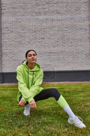 stylische Sportlerin in lindfarbenem Kapuzenpulli und Leggings auf grünem Gras sitzend, gesunder Lebensstil