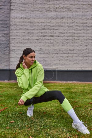attraktive Sportlerin in lindfarbenem Kapuzenpulli und Leggings auf grünem Gras sitzend, gesunder Lebensstil