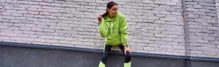 attractive sportswoman in hoodie and leggings touching ponytail and standing near grey wall, banner