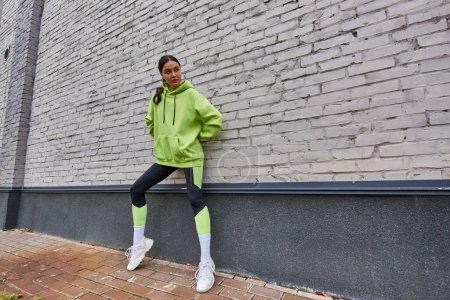 stylish young sportswoman in lime color hoodie, leggings and sneakers standing near grey wall