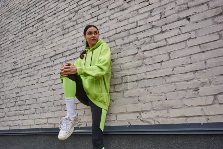 stylische junge Sportlerin in lindfarbenem Kapuzenpulli und Leggings, die in der Nähe der grauen Wand im Freien trainiert