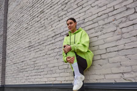 stylish young sportswoman in lime color hoodie and leggings stretching near grey wall outdoors