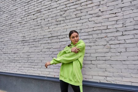 attractive young sportswoman in lime color hoodie and leggings stretching near grey wall outdoors