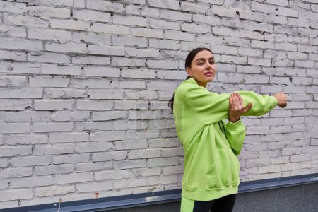 motivated young sportswoman in lime color hoodie and leggings stretching near grey wall outdoors