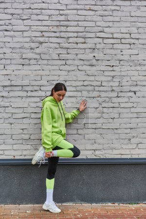 pretty young sportswoman in lime color hoodie and leggings stretching leg near grey wall outdoors