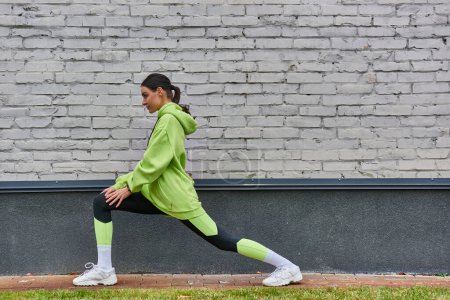 pretty young sportswoman in lime color hoodie and leggings doing lunges before running outdoors