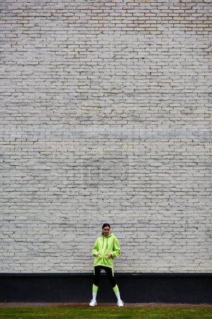 Foto de Bonita deportista en sudadera con capucha color lima y leggings de pie con las manos en los bolsillos cerca de la pared de ladrillo - Imagen libre de derechos