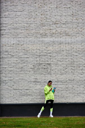 Glückliche Sportlerin in Kapuzenpulli und Leggings mit Smartphone und Wasserflasche