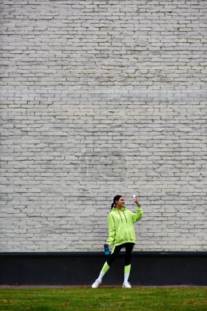 glückliche Sportlerin in Kapuzenpulli und Leggings, die Foto auf dem Smartphone spricht und Flasche mit Wasser hält