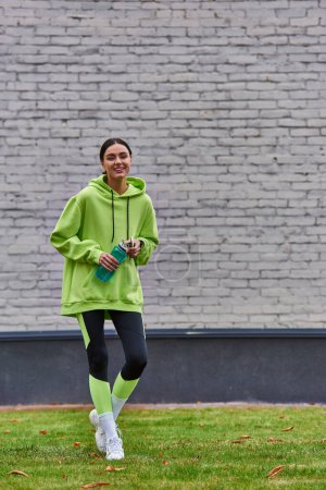 Positive Sportlerin in Kapuzenpulli und Leggings mit Smartphone und Wasserflasche