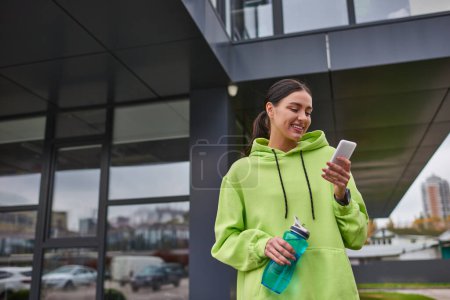 Glückliche Sportlerin im kuscheligen Kapuzenpulli mit Smartphone und mit Flasche mit Wasser im Freien