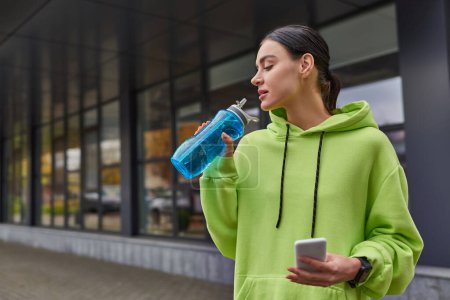 Sportlerin in lindfarbenem Kapuzenpulli hält Smartphone und trinkt nach dem Training Wasser aus Flasche
