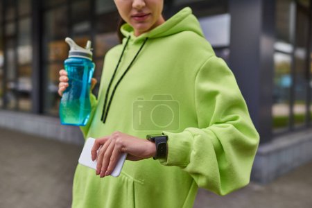 deportista recortado sosteniendo el teléfono inteligente y la botella mientras revisa el rastreador de fitness después del entrenamiento