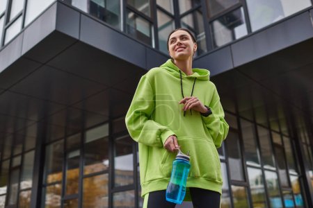 Glückliche Sportlerin in überdimensionalem Kapuzenpulli und Leggings, die mit Flasche mit Wasser im Freien steht